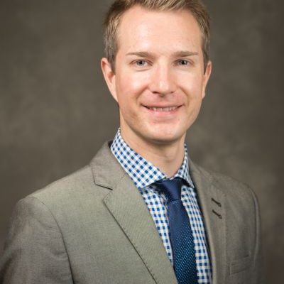 Alexy is blonde and is wearing a grey suit with a blue and white shirt and a blue tie