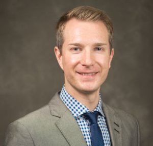 Alexy is blonde and is wearing a grey suit with a blue and white shirt and a blue tie