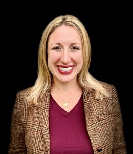 Bekah on a black background with blonde hair, a maroon shirt, plaid brown jacket