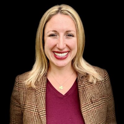 Bekah on a black background with blonde hair, a maroon shirt, plaid brown jacket