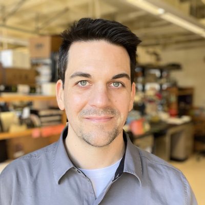 He has dark hair, blue eyes, and is wearing a grey, blue business shirt. He has a lab in the background.