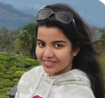 She is in a sweatshirt and sunglasses and has dark, long hair. The background is a sky with mountains and greenery.