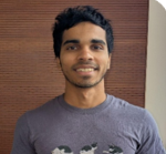 He is in a grey t shirt with a brown background. He has dark, short hair. 