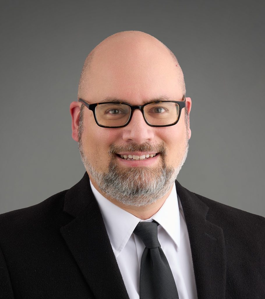 Christian in a black suit and tie with a white undershirt and glasses