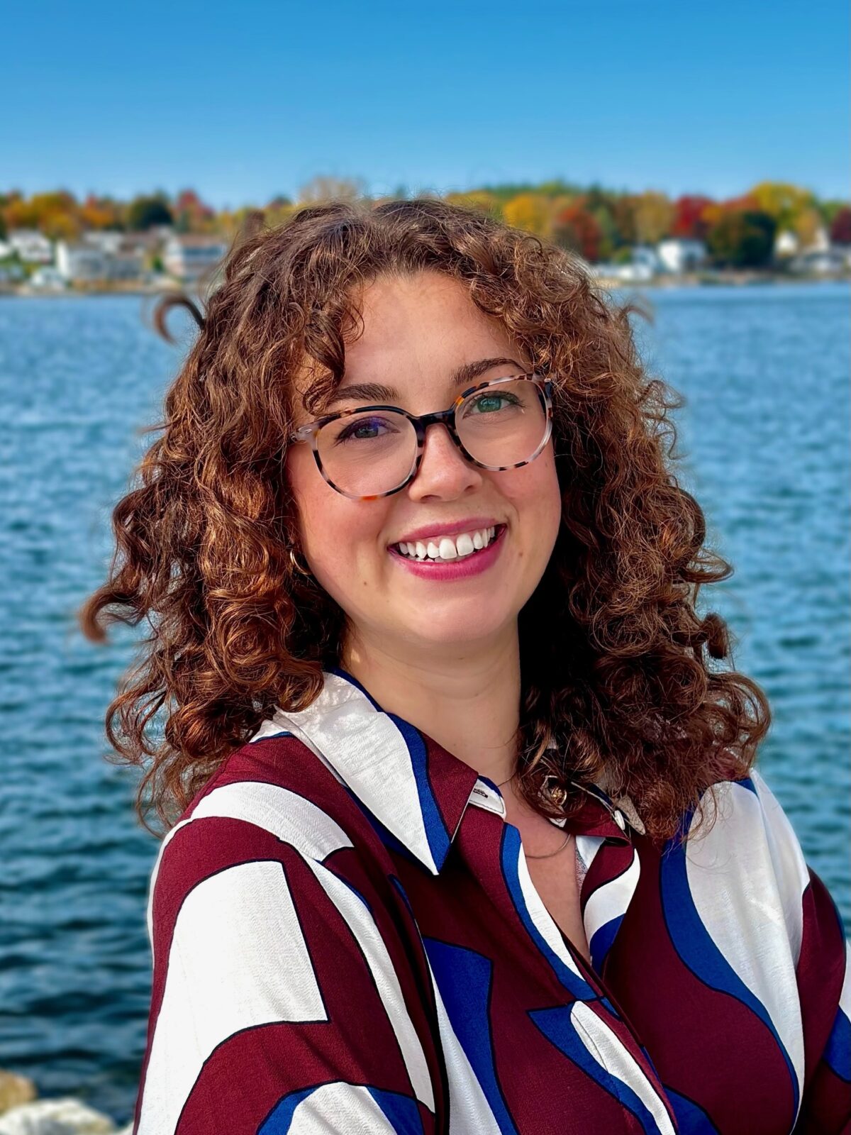 Portia in a blue, maroon, and white shirt. She has curly hair and glasses. She is standing in front of the water. 