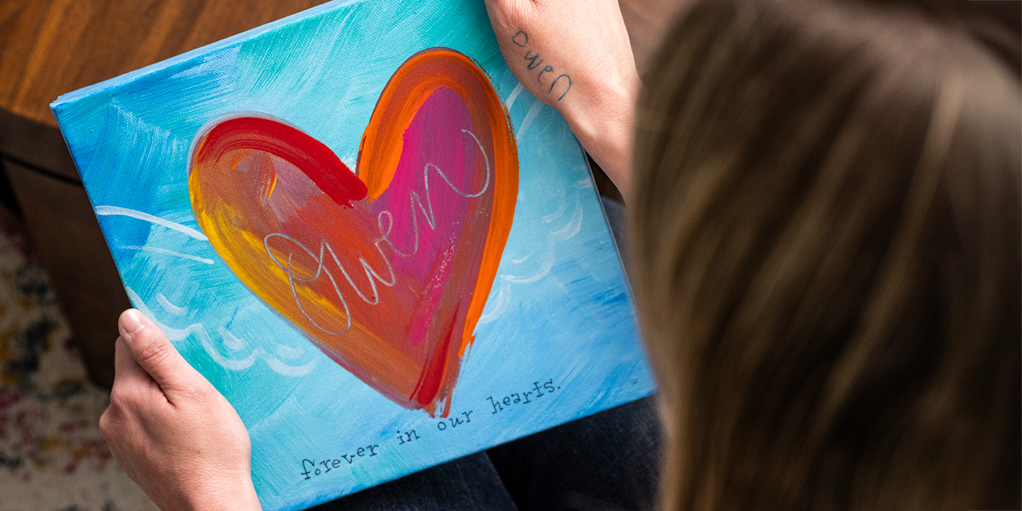 women with brown hair holds a heart image and shows her tattoo of her son's name on her hand